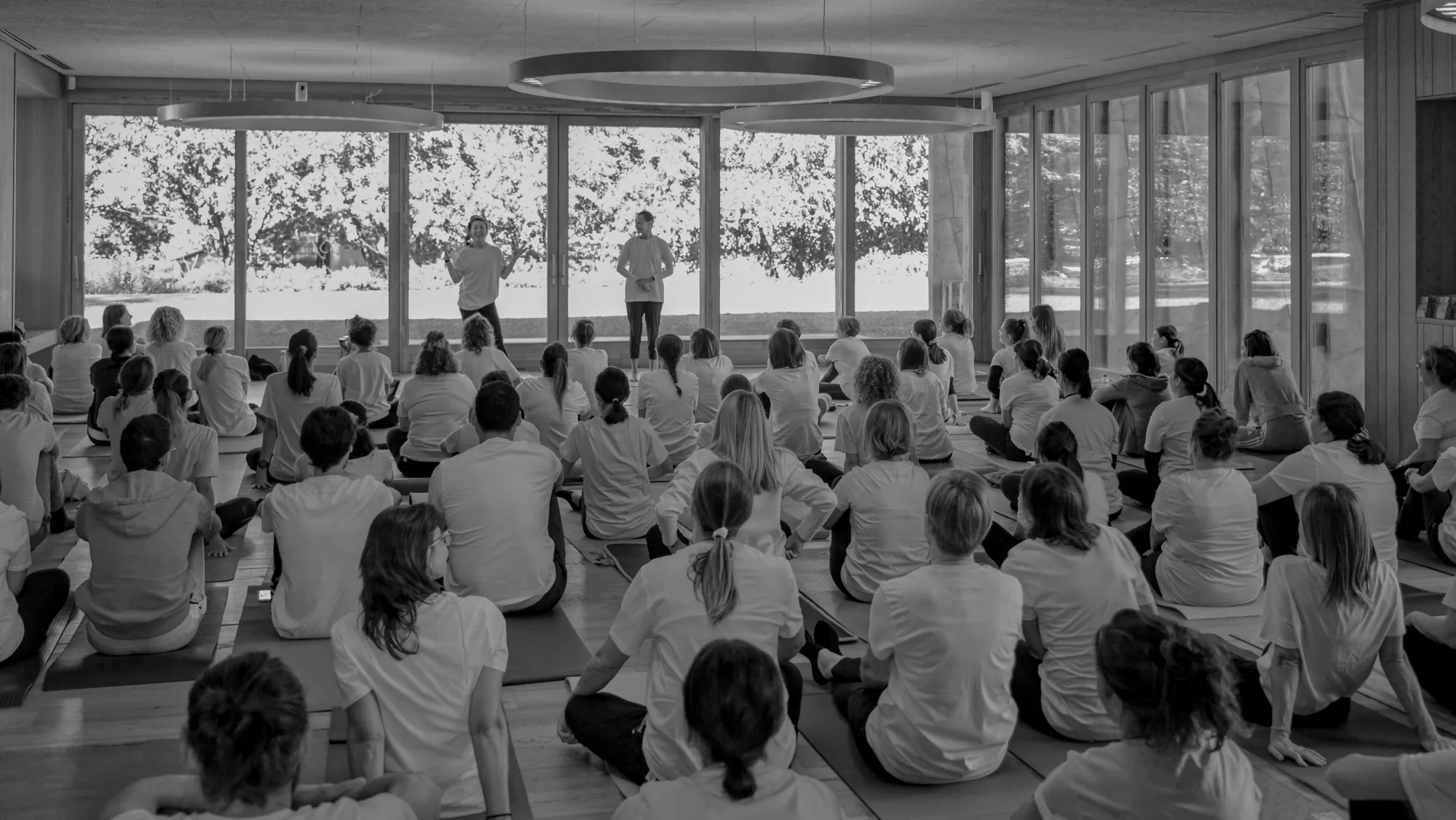 The full hall at the first edition of Yoga per la Ricerca: participants on their mats listen to the two teachers in front of the windows
